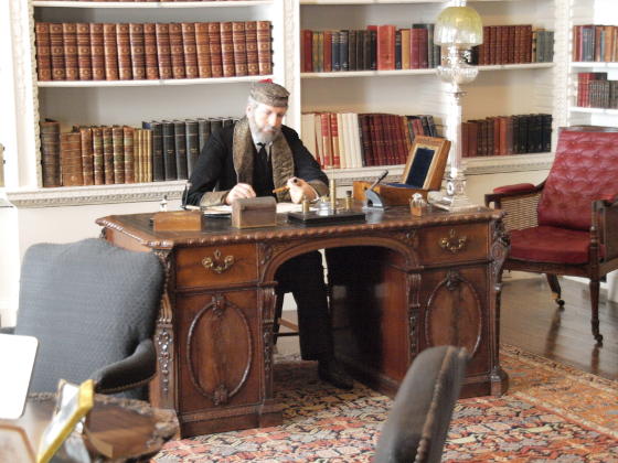 A waxwork of a man in a smoking cap and jacket sitting at a desk in a library
